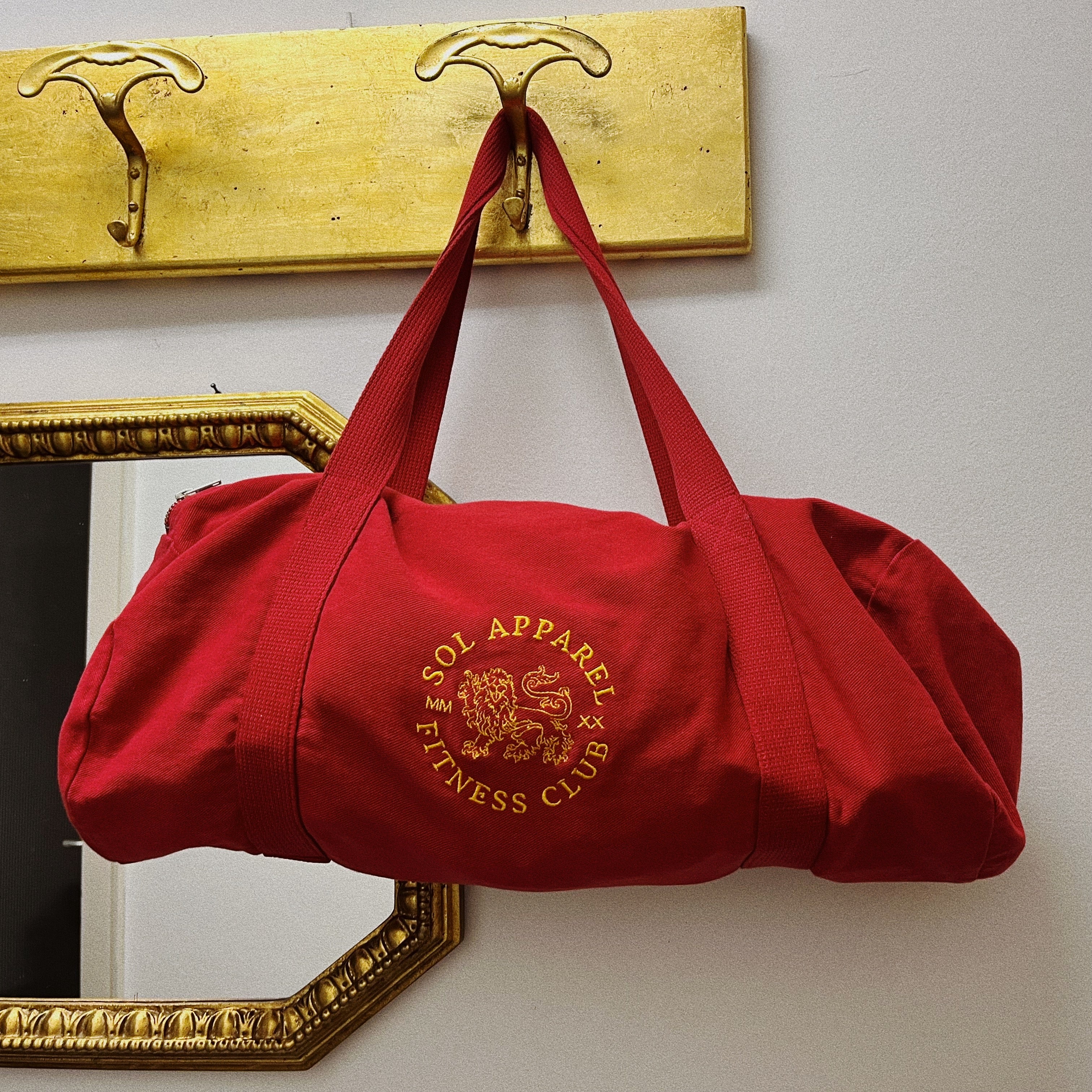 red gym duffel bag with golden logo of lion embroidered hanging from a hook in a house with white walls