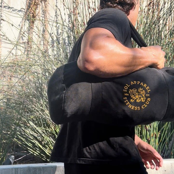 man walking past a wooden fence with a black cotton tshirt and a natural cotton duffel bag in black with a golden embroidered lion logo