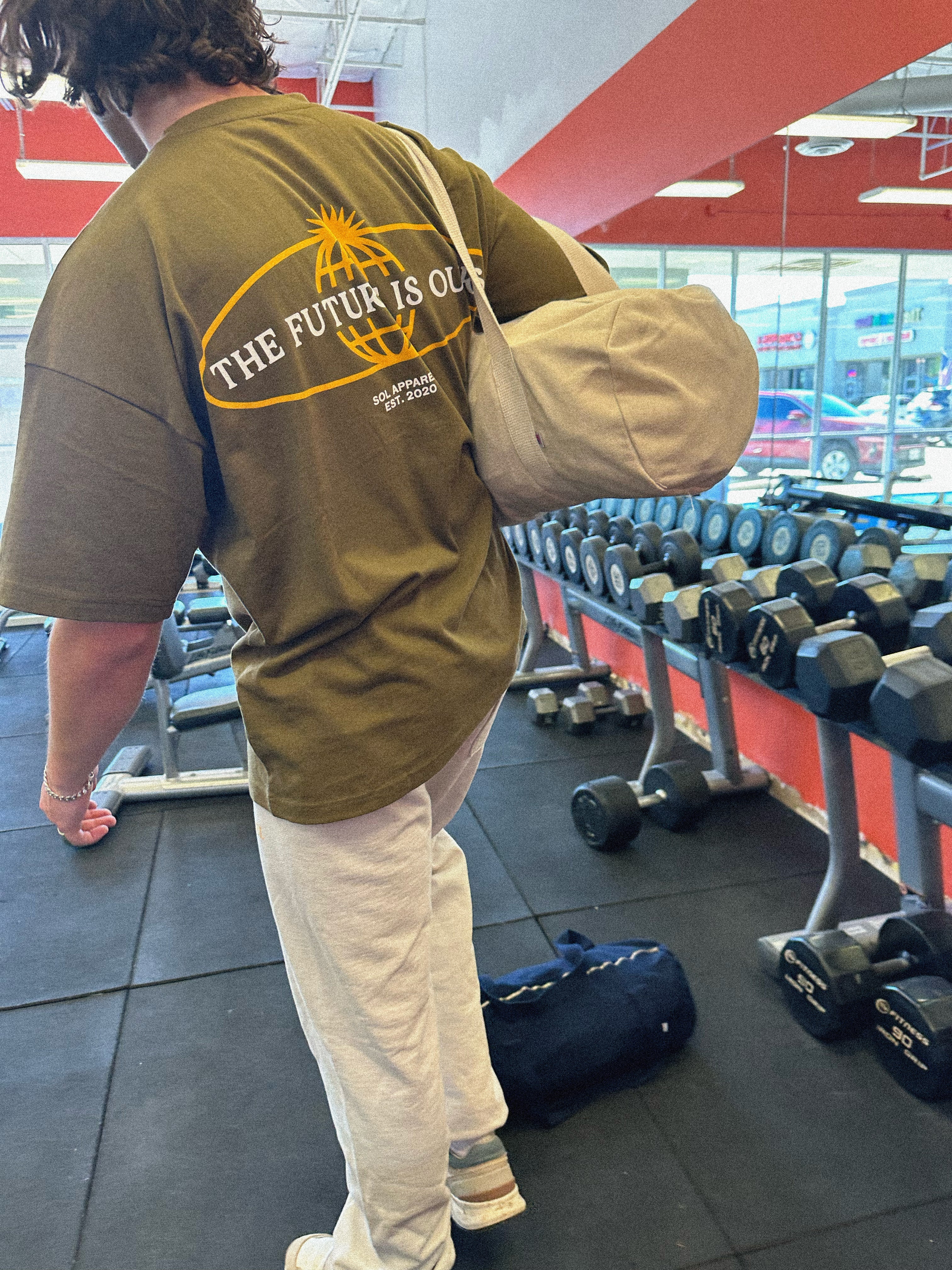 man in gym with oversized olive green cotton tshirt that says the future is ours in white writing