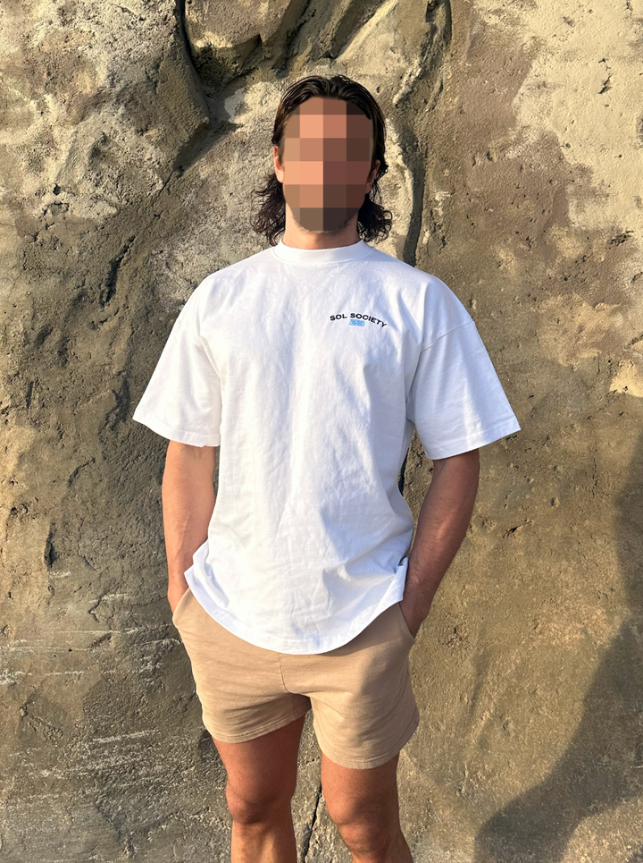 sol brah standing in front of rocks at the beach with an oversized white retro cotton gym tee that says sol society on the left chest